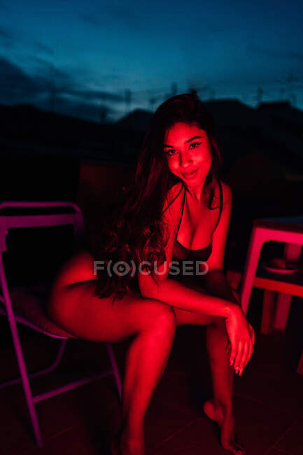 Молода жінка в нижній білизні дивиться на камеру, сидячи на стільці під червоним неоновим світлом вночі на терасі — стокове фото