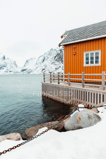 Cottage giallo e molo innevato situato vicino al mare increspato contro le montagne nella fredda giornata invernale nel villaggio costiero sulle isole Lofoten, Norvegia — Foto stock