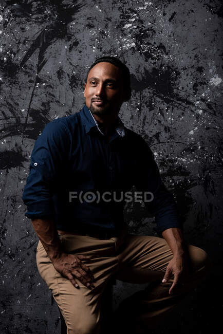 Ernster, stilvoller ethnischer Mann sitzt auf einem Holzhocker im Studio auf dunklem Hintergrund und schaut weg — Stockfoto