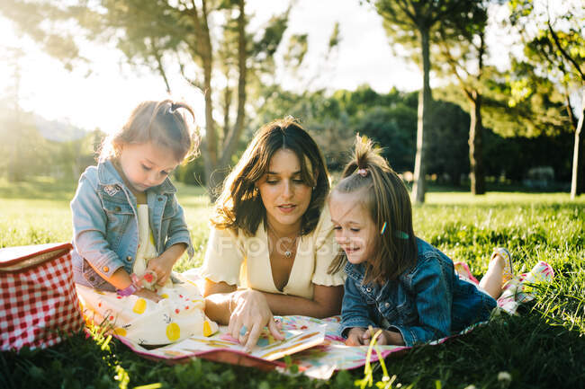Щаслива молода мама з маленькими доньками лежить на ковдрі і грає в гру, проводячи літній день разом у сонячному парку — стокове фото