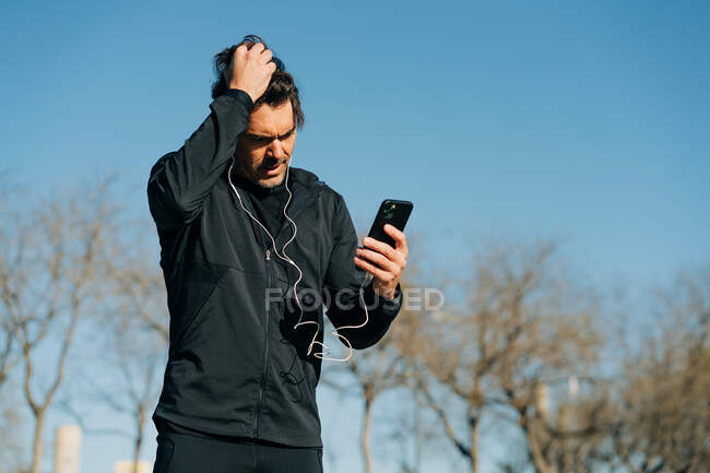 Bearded male athlete in sportswear and earphones browsing cellphone while standing on meadow in town — Stock Photo