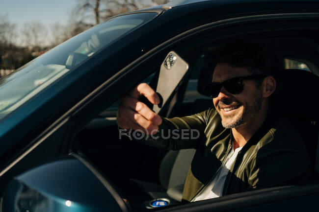 Happy male in sunglasses taking selfie on cellphone while sitting in automobile on sunny day — Stock Photo