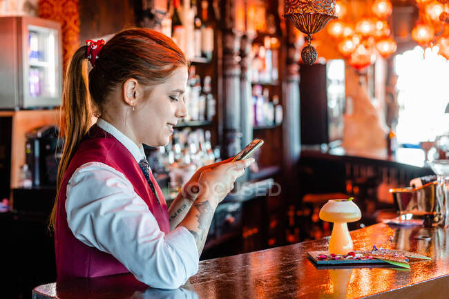 Vista lateral de garçom feminino alegre tirando foto de coquetel azedo em vidro servido no balcão no pub — Fotografia de Stock