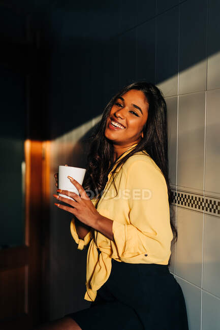 Vista lateral da jovem alegre fêmea hispânica em camisa amarela desfrutando de bebida quente enquanto se inclina na parede e descansa de manhã em casa olhando para a câmera — Fotografia de Stock
