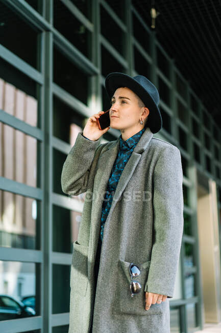 Vista lateral de la persona andrógina en sombrero hablando en el teléfono celular mientras mira hacia otro lado de pie en la calle a la luz del día - foto de stock