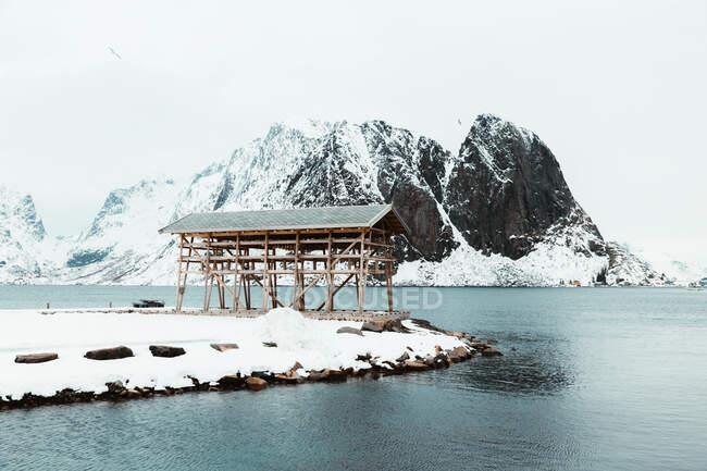 Estructura de madera situada en la costa nevada cerca del mar contra la cresta de la montaña y el cielo gris en el frío día de invierno en las islas Lofoten, Noruega - foto de stock