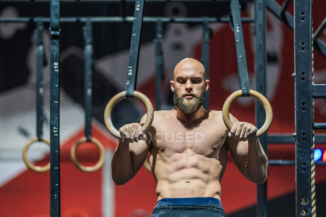 Сильний без сорочки чоловік дивиться вниз стоячи вправу на гімнастичних кільцях під час інтенсивного тренування в сучасному тренажерному залі — стокове фото