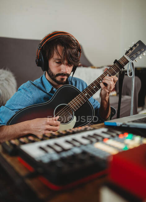 Hombre joven en auriculares tocando en la guitarra cerca de la mesa con el ordenador portátil y sintetizador en casa mirando a la cámara - foto de stock