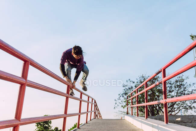 Low angle fearless young man jumping above metal railing in city while performing parkour stunt on sunny day — Stock Photo