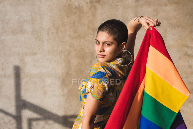 Grave giovane donna etnica bisessuale con bandiera multicolore che rappresenta i simboli LGBTQ e guarda la fotocamera nella giornata di sole — Foto stock