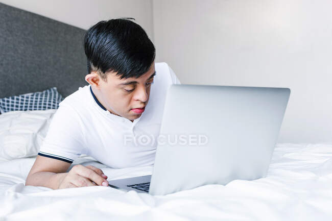 Focado Latina adolescente menino com síndrome de Down deitado na cama e navegar na Internet no netbook — Fotografia de Stock