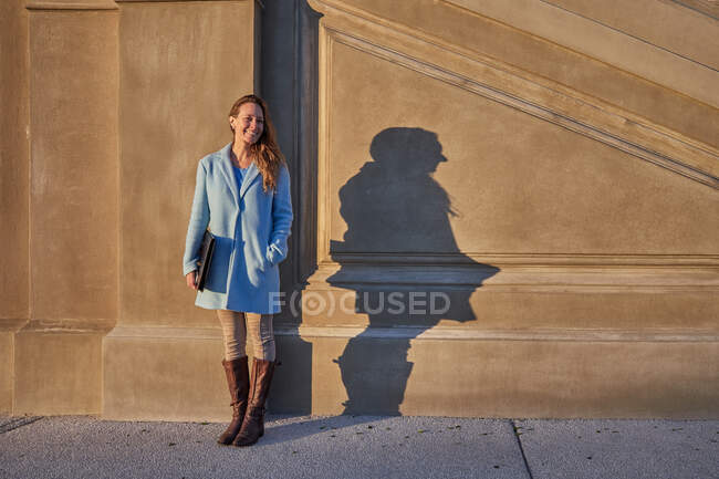 Full body of happy adult female in blue coat standing near stone building wall with folder in hands in city street in sunny day — Stock Photo