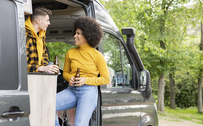 Mujer negra feliz con la cerveza sonriendo mirándose el uno al otro de pie en furgoneta con novio durante el viaje por carretera en el campo - foto de stock