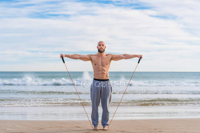 Hombre brutal sin camisa mirando a la cámara haciendo moscas laterales laterales con entrenamiento de banda resistente en la orilla del mar y mirando hacia adelante - foto de stock