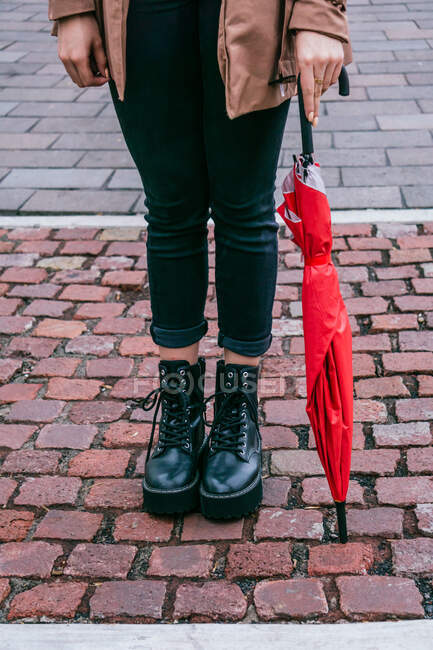 Femme anonyme en vêtements décontractés et avec parapluie debout sur le trottoir pavé dans la rue de la ville — Photo de stock