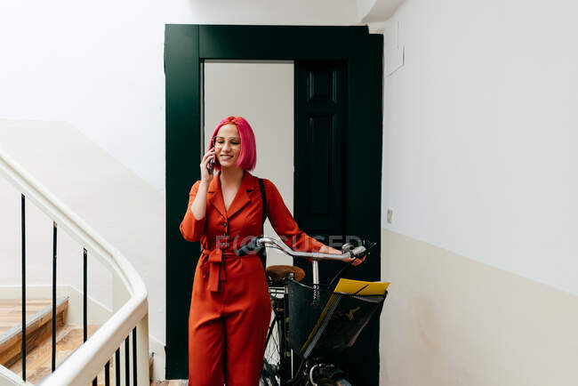 Young stylish female in red suit with backpack talking on smartphone while standing with bicycle on staircase — Stock Photo