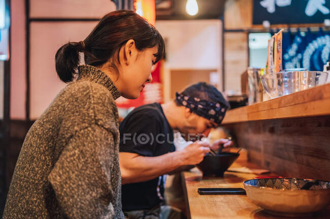 Vista lateral da mulher e do homem se comunicando enquanto come comida asiática no balcão de madeira no café moderno — Fotografia de Stock