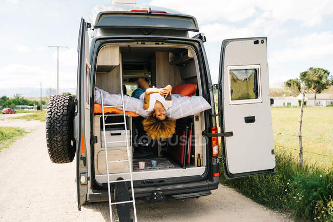 Positive black woman with Afro hairstyle lying upside down on bed in trailer and browsing mobile phone on sunny summer day — Stock Photo