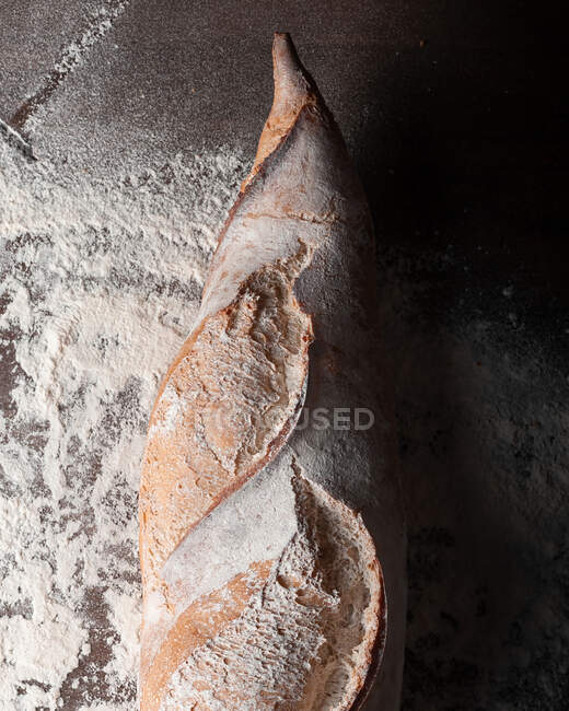 Apetitiva baguette recién horneada con corteza crujiente colocada sobre una mesa de madera cubierta con harina blanca sobre fondo negro - foto de stock