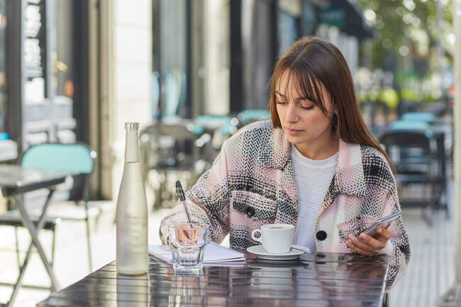 Young woman taking notes in notebook while browsing on smartphone sitting at table in outdoor cafe in city — Stock Photo