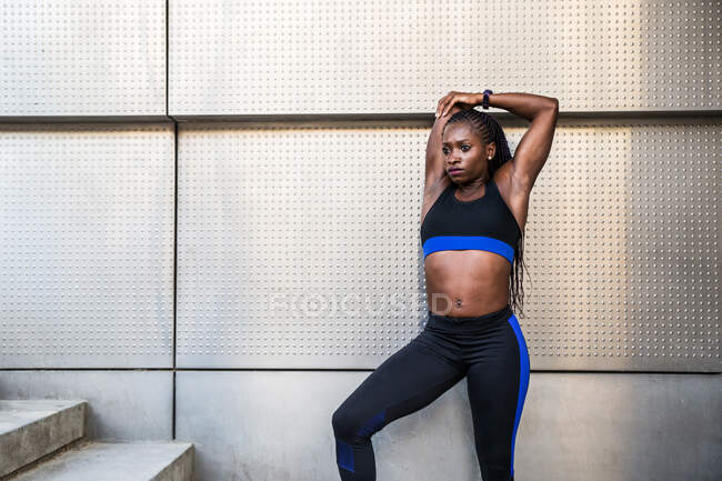 Mujer bastante negra en ropa deportiva manteniendo los ojos cerrados y haciendo ejercicios de calentamiento cerca de la pared del edificio moderno en la calle de la ciudad - foto de stock