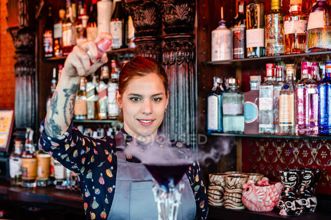 Entzückte Barkeeperin in Schürze bereitet Aroma-Blaster-Cocktail an Holztheke in Bar zu — Stockfoto