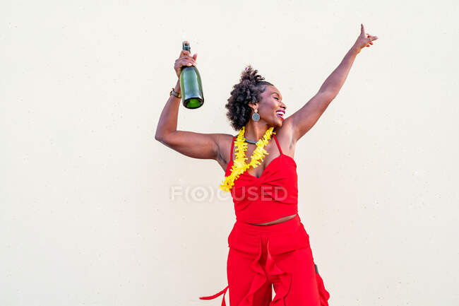 Mulher afro-americana encantada em colar de flores vestindo brilhante posição geral com garrafa de champanhe contra fundo branco — Fotografia de Stock