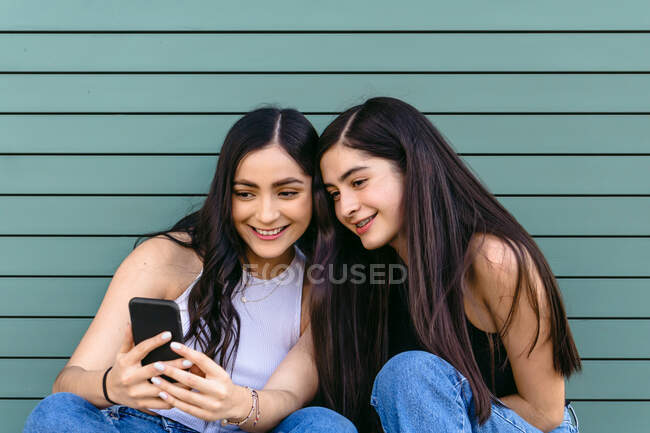 Zufriedene weibliche Geschwister mit langen Haaren surfen mit dem Handy im Internet, während sie bei Tageslicht Zeit miteinander verbringen — Stockfoto