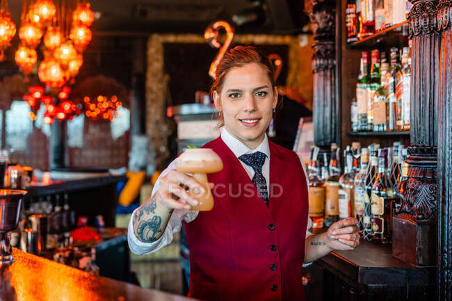 Cantinera sonriente parada en el mostrador del bar con un tipo de bebida alcohólica servida en vasos de cóctel creativos en forma de champiñón - foto de stock