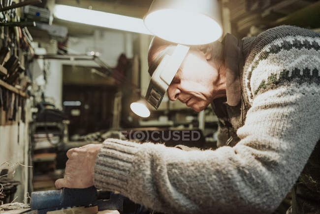 Side view of focused senior luthier in sweater and magnifier head strap standing at workbench while repairing string instrument detail in old studio — стоковое фото