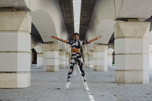Young fitness African American woman making exercise jumping and listening music after running on city street — Stock Photo