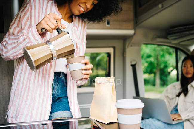 Crop positive young African American woman pouring coffee from kettle into cup while resting with Asian girlfriend inside camper van during summer journey in nature — Stock Photo