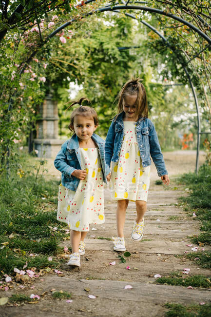 Full body of adorable little girls in similar dresses and denim jackets holding hands while walking together through arched pathway in green summer park — Stock Photo