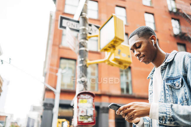 De baixo conteúdo alegre afro-americano cara em jeans jaqueta na moda surfar telefone celular moderno durante o passeio pela cidade — Fotografia de Stock