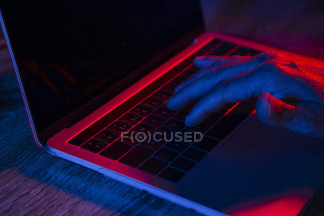 Crop anonymous male using laptop with black screen placed on wooden table in dark workplace with neon lights — Stock Photo