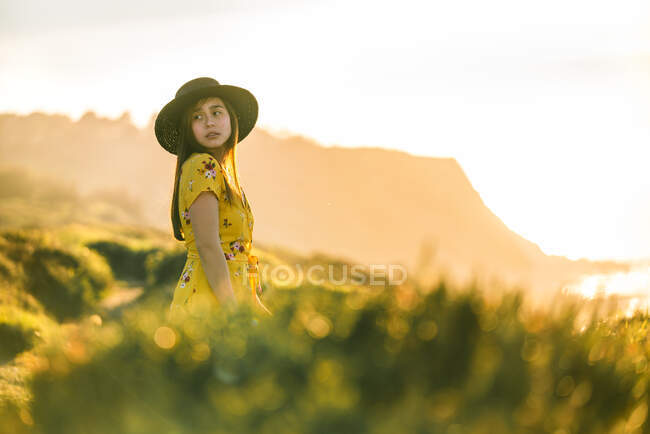 Attraente giovane femmina in giallo prendisole e cappello in piedi su verdeggiante prato erboso in campagna soleggiata — Foto stock