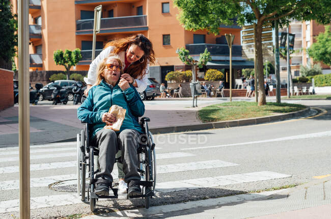 Adult woman pushing wheelchair with senior mother and crossing road in city during stroll in summer — Stock Photo