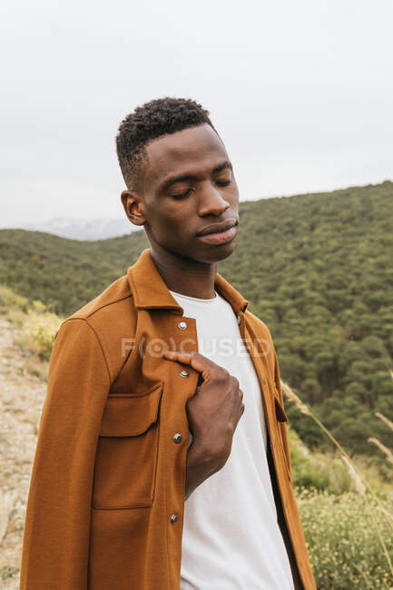 Hombre afroamericano pensativo en ropa de moda con los ojos cerrados en la naturaleza y admirando la vista del bosque montañoso - foto de stock