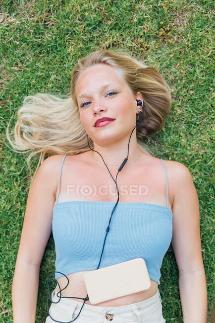 Visão superior da fêmea sonhadora desfrutando de músicas em fones de ouvido enquanto estava deitada olhando para a câmera na grama no parque no verão — Fotografia de Stock
