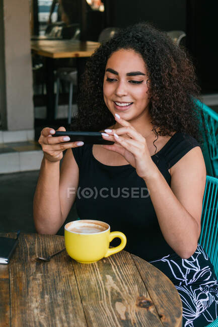 Joven mujer hispana de pelo rizado positiva tomando una foto de una taza de capuchino aromático fresco mientras pasa tiempo libre en la terraza de la cafetería - foto de stock