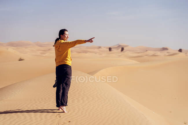 Увага до усмішки молодої босоногої жінки в повсякденному одязі, що стоїть на піщаній дюні проти пустелі і вказує на подорож в Емірати. — стокове фото