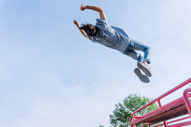 De baixo de bravo macho pulando sobre cerca de metal na rua e mostrando truque parkour contra o céu azul — Fotografia de Stock