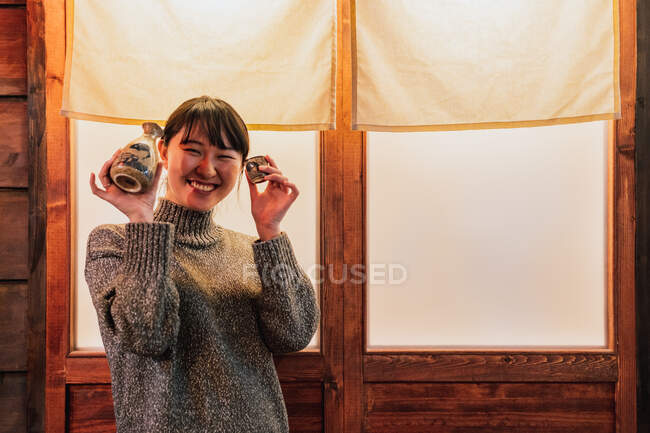 Mulher asiática em suéter de pé perto da porta com bebida tradicional no café — Fotografia de Stock