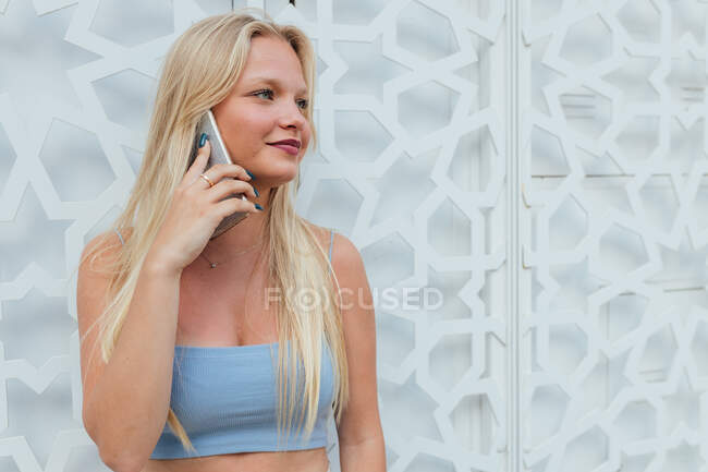 Спокійна усміхнена жінка з світлим волоссям і в літньому вбранні, стоячи в місті і розмовляючи на мобільному телефоні, дивлячись вбік — стокове фото