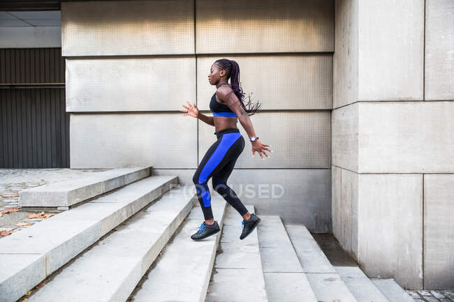 Вид сбоку на сильную афроамериканку, бегущую по лестнице во время тренировки возле современного здания на городской улице — стоковое фото