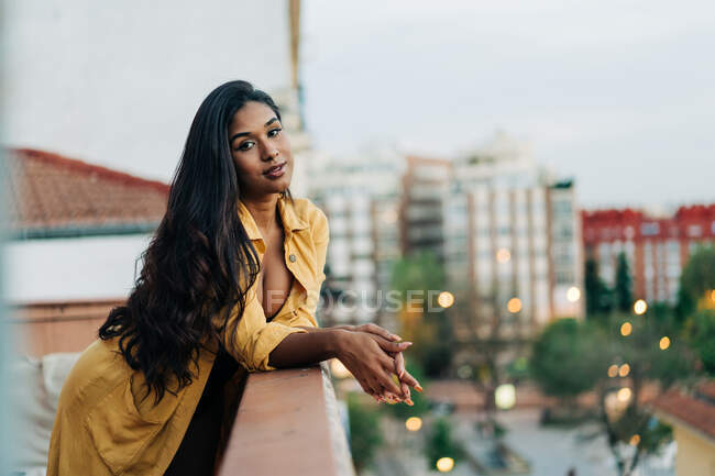 Young Hispanic female in casual clothes leaning on railing and looking at camera while relaxing on balcony in evening in city — Stock Photo