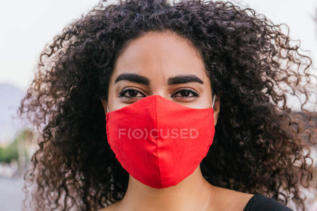 Young curly haired Hispanic female on red fabric protective mask for coronavirus prevention while spending summer day on street looking at camera — Stock Photo