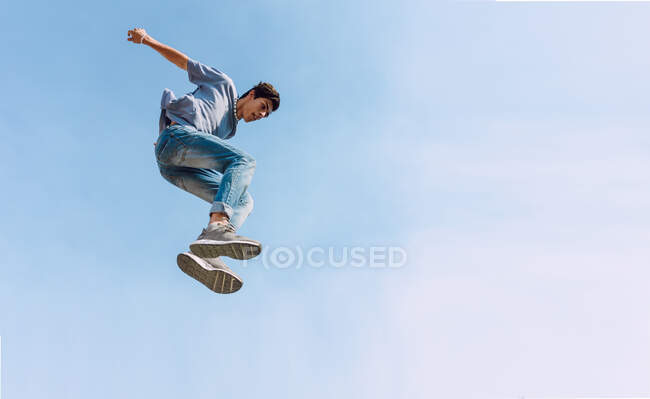 Desde abajo del macho saltando por encima del suelo y realizando acrobacias de parkour sobre el fondo del cielo azul sin nubes - foto de stock