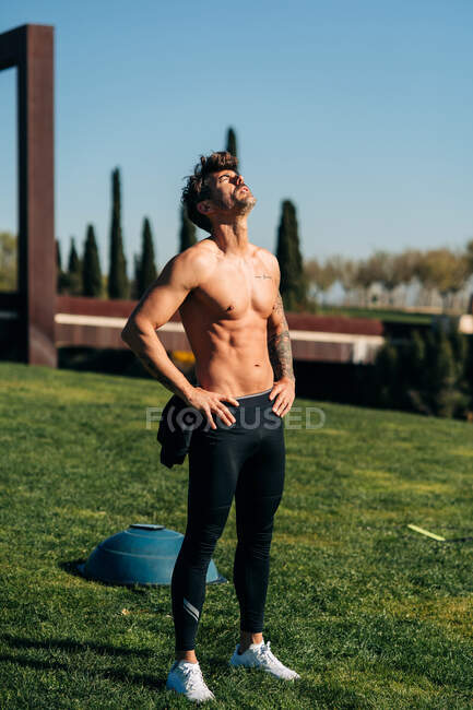 Muscular male athlete with naked torso and hands on hips having break from training on meadow — Stock Photo