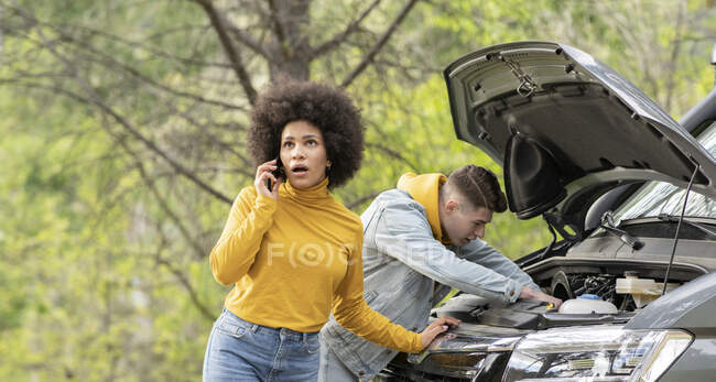 Schockierte schwarze Frau ruft Rettungsdienst an, während junger Mann versucht, Motor von Transporter während einer Autofahrt auf dem Land zu reparieren — Stockfoto